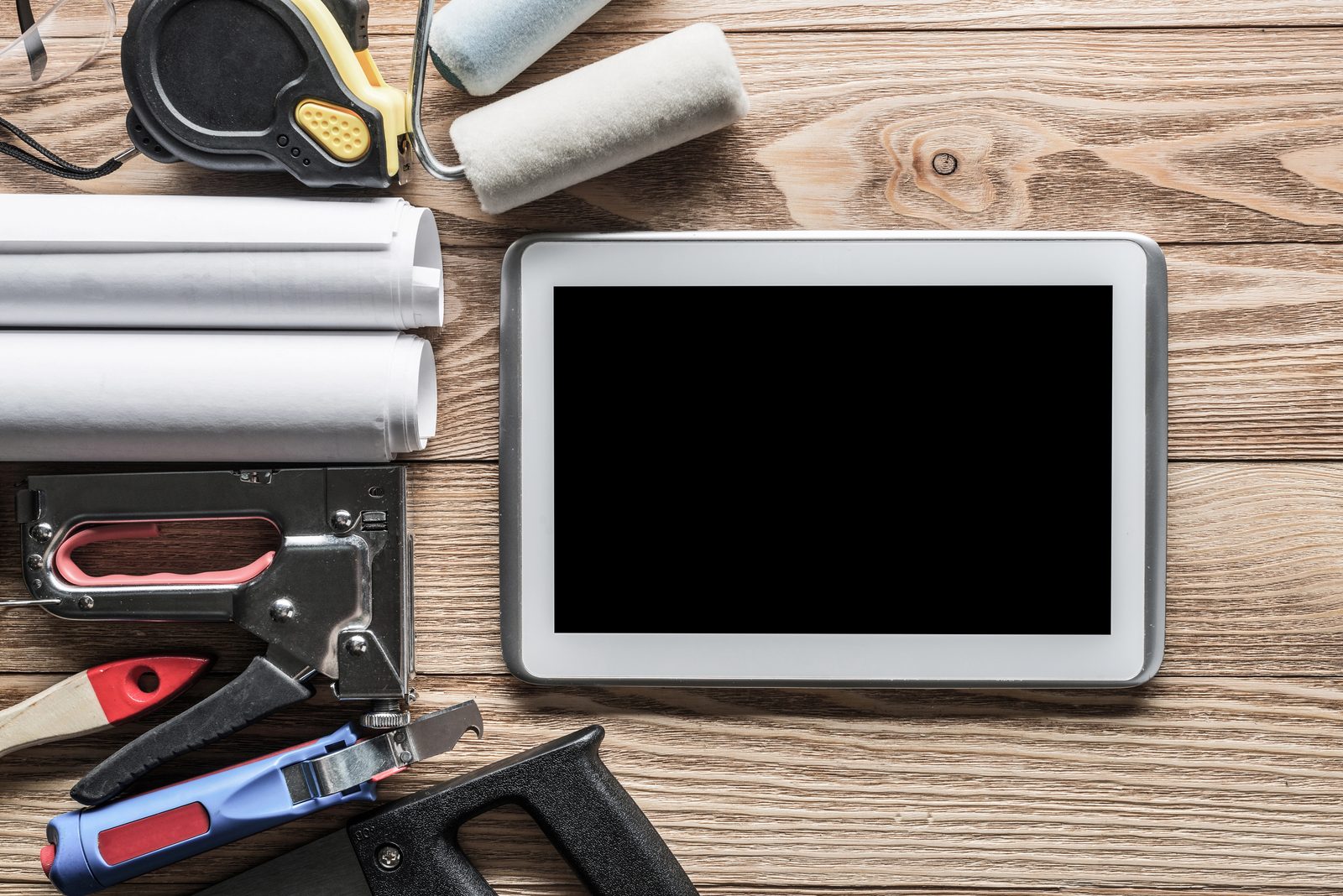 Tablet neben Werkzeugen, Bauplänen und Farbrolle auf einer Holzwerkbank