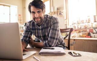 Handwerker sitzt lächelnd mit Laptop am Tisch in seiner Werkstatt