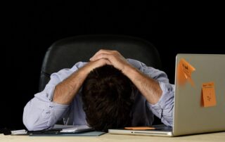 Mann sitzt erschöpft am Schreibtisch und hält die Hände über den Kopf